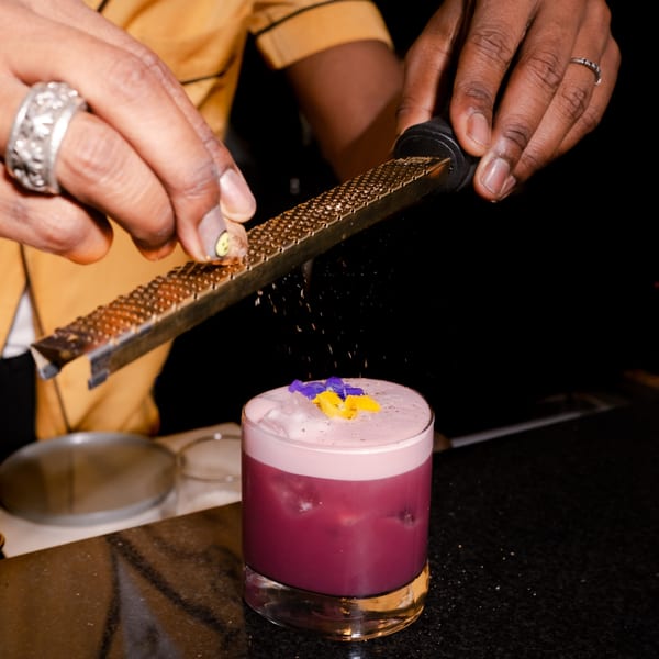 A bartender grates a garnish over a vibrant purple cocktail topped with foam and edible flowers.