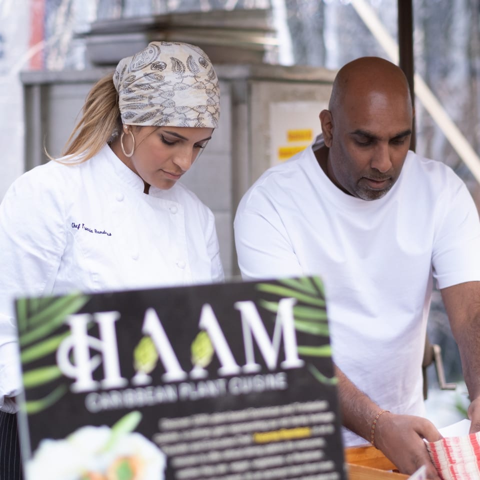 Two chefs, a woman and a man, working behind a HAAM Caribbean Plant Cuisine sign.