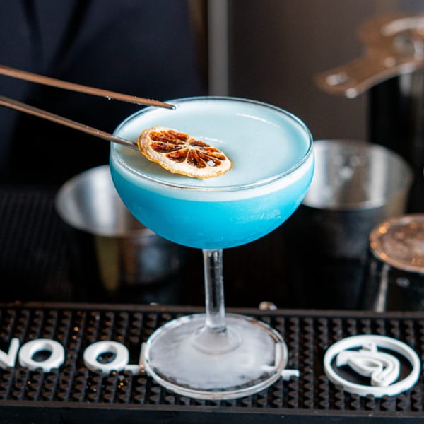A vibrant blue cocktail in a coupe glass, garnished with a dried orange slice, on a bar.